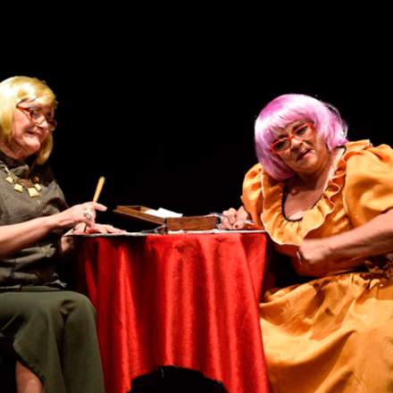 Dos artistas disfrazados de personas mayores sentadas en una mesa